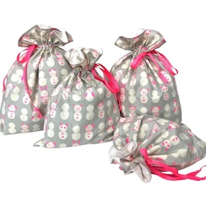 May include: Four gray fabric gift bags with a repeating pattern of white snowmen with pink accents. Each bag is cinched closed with a bright pink ribbon. The bags are of varying sizes.