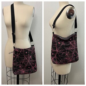 K&ouml;nnte beinhalten: Eine schwarze Umh&auml;ngetasche mit einem pinkfarbenen Spinnennetz-Muster und kleinen pinkfarbenen Spinnen. Die Tasche hat einen schwarzen verstellbaren Riemen und einen silbernen Ringverschluss.