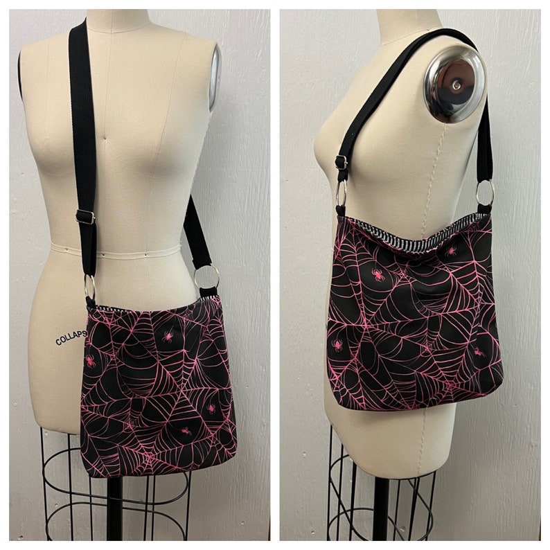 K&ouml;nnte beinhalten: Eine schwarze Umh&auml;ngetasche mit einem pinkfarbenen Spinnennetz-Muster und kleinen pinkfarbenen Spinnen. Die Tasche hat einen schwarzen verstellbaren Riemen und einen silbernen Ringverschluss.