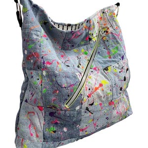 May include: A hobo-style shoulder bag constructed from patchwork denim fabric featuring a vibrant neon paint splatter pattern in pink, green, yellow, and orange. The bag includes a diagonal zippered front pocket and black strap attachments.