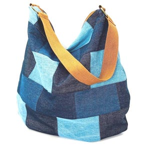 May include: A patchwork denim shoulder bag in shades of blue and light blue. The bag features a wide, tan-colored strap and a spacious design. The denim is sewn together in a patchwork pattern.