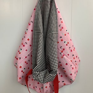 K&ouml;nnte beinhalten: Eine rosa Tragetasche mit Kirschendruck und roten Griffen. Die Tasche ist reversibel und hat auf der anderen Seite ein schwarz-wei&szlig;es Gingham-Muster.
