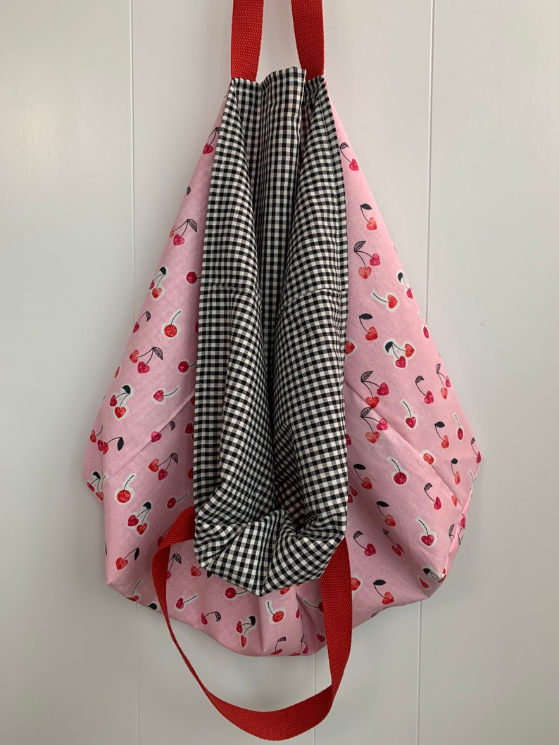 K&ouml;nnte beinhalten: Eine rosa Tragetasche mit Kirschendruck und roten Griffen. Die Tasche ist reversibel und hat auf der anderen Seite ein schwarz-wei&szlig;es Gingham-Muster.