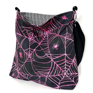 K&ouml;nnte beinhalten: Schwarze Canvas-Tragetasche mit einem pinkfarbenen Spinnennetz-Muster und pinkfarbenen Spinnen. Die Tasche hat einen schwarzen verstellbaren Riemen.