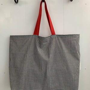 K&ouml;nnte beinhalten: Eine schwarze und wei&szlig;e Gingham-Tragetasche mit roten Griffen. Die Tasche h&auml;ngt an Haken an einer wei&szlig;en Wand.