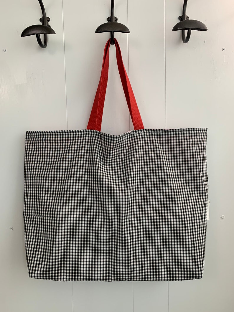 K&ouml;nnte beinhalten: Eine schwarze und wei&szlig;e Gingham-Tragetasche mit roten Griffen. Die Tasche h&auml;ngt an Haken an einer wei&szlig;en Wand.