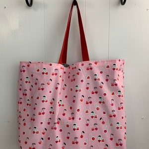 K&ouml;nnte beinhalten: Eine rosa Canvas-Tragetasche mit roten Griffen und einem Kirschendruck. Die Tasche h&auml;ngt an Haken an einer wei&szlig;en Wand.