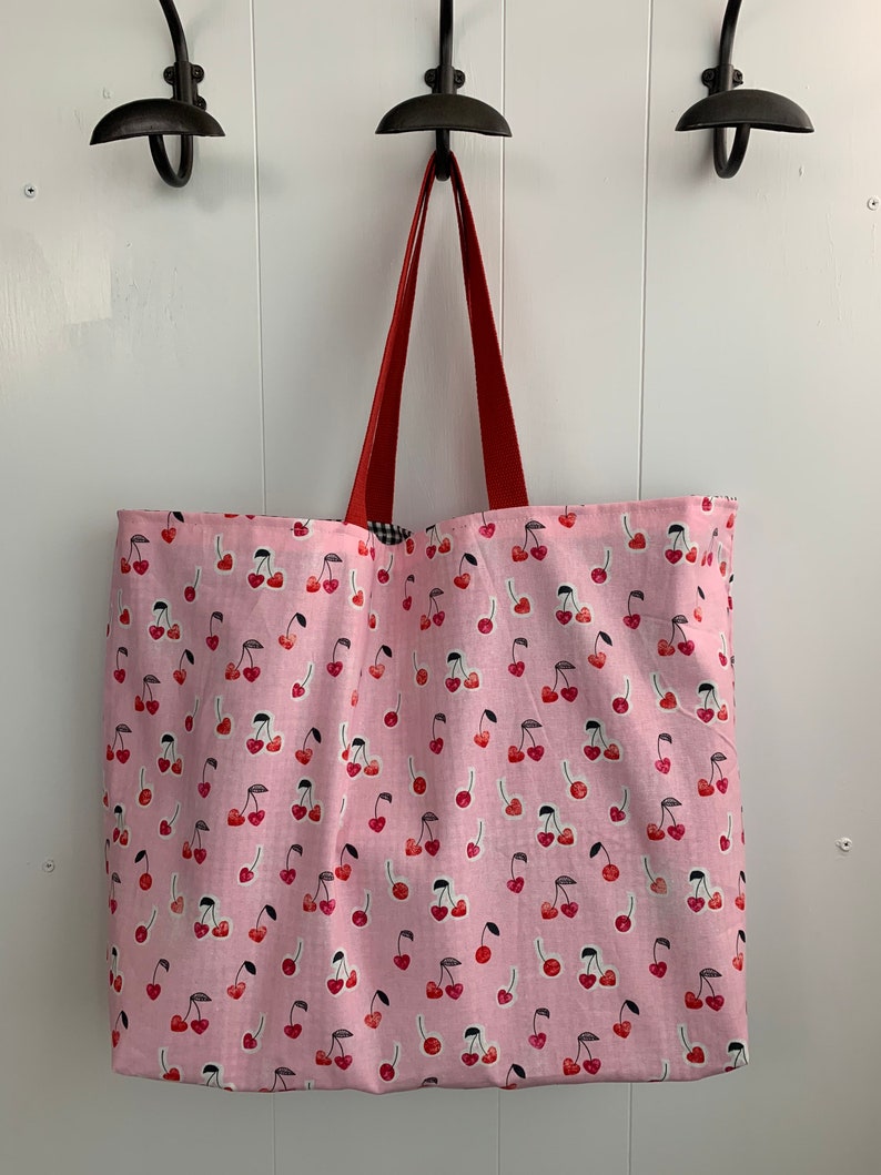 K&ouml;nnte beinhalten: Eine rosa Canvas-Tragetasche mit roten Griffen und einem Kirschendruck. Die Tasche h&auml;ngt an Haken an einer wei&szlig;en Wand.