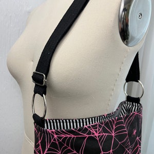 K&ouml;nnte beinhalten: Eine schwarze Schultertasche mit einem pinkfarbenen Spinnennetz-Muster und pinkfarbenen Spinnen. Die Tasche hat einen schwarzen verstellbaren Riemen mit silbernen Ringen.
