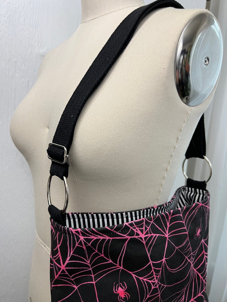 K&ouml;nnte beinhalten: Eine schwarze Schultertasche mit einem pinkfarbenen Spinnennetz-Muster und pinkfarbenen Spinnen. Die Tasche hat einen schwarzen verstellbaren Riemen mit silbernen Ringen.