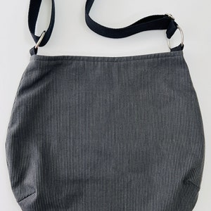 May include: A dark gray and white striped shoulder bag with a black strap and silver metal rings. The bag is made of a soft fabric and has a rounded shape.