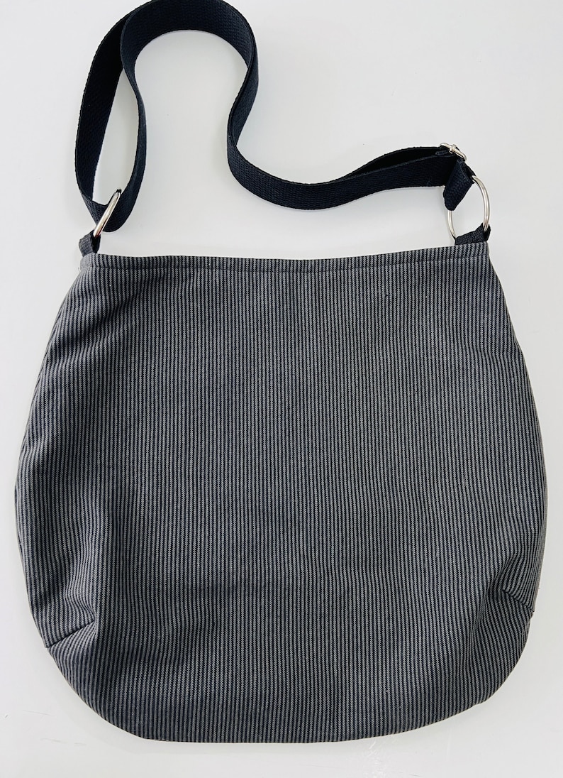 May include: A dark gray and white striped shoulder bag with a black strap and silver metal rings. The bag is made of a soft fabric and has a rounded shape.