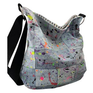 May include: A hobo bag constructed from light blue denim patches featuring a vibrant paint splatter pattern in neon pink, green, orange, and black. The bag includes a wide black fabric shoulder strap, silver metal rings, and a black and white striped lining.