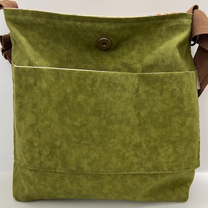 Puede incluir: Un bolso bandolera de lona verde con una correa marr&oacute;n y un cierre de presi&oacute;n &uacute;nico. El bolso tiene un bolsillo grande en la parte delantera.
