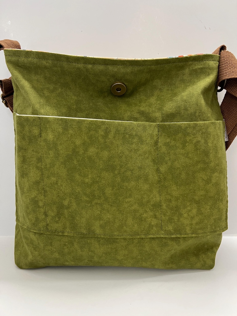 Puede incluir: Un bolso bandolera de lona verde con una correa marr&oacute;n y un cierre de presi&oacute;n &uacute;nico. El bolso tiene un bolsillo grande en la parte delantera.