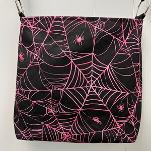 K&ouml;nnte beinhalten: Schwarze Stofftasche mit einem pinkfarbenen Spinnennetz-Muster. Die Tasche hat in jeder Ecke eine pinkfarbene Spinne.