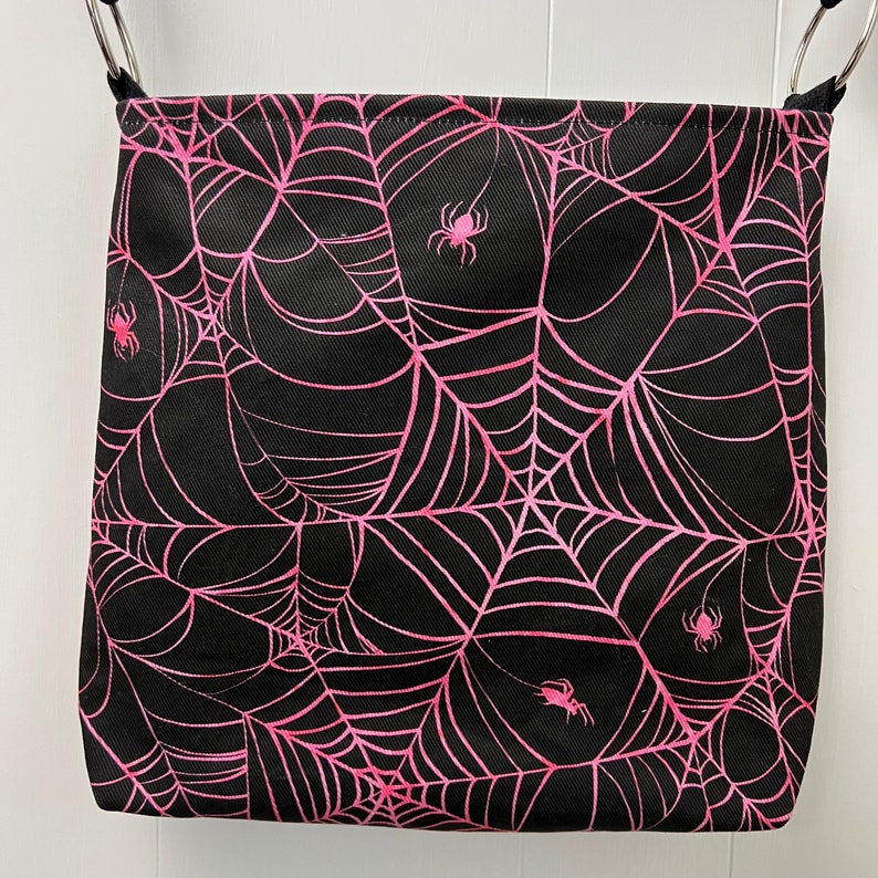 K&ouml;nnte beinhalten: Schwarze Stofftasche mit einem pinkfarbenen Spinnennetz-Muster. Die Tasche hat in jeder Ecke eine pinkfarbene Spinne.