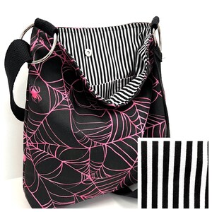 K&ouml;nnte beinhalten: Schwarze Canvas-Schultertasche mit einem pinkfarbenen Spinnennetz-Design. Die Tasche hat ein schwarz-wei&szlig; gestreiftes Futter.