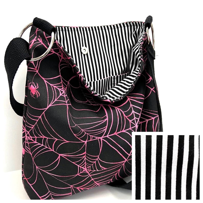 K&ouml;nnte beinhalten: Schwarze Canvas-Schultertasche mit einem pinkfarbenen Spinnennetz-Design. Die Tasche hat ein schwarz-wei&szlig; gestreiftes Futter.