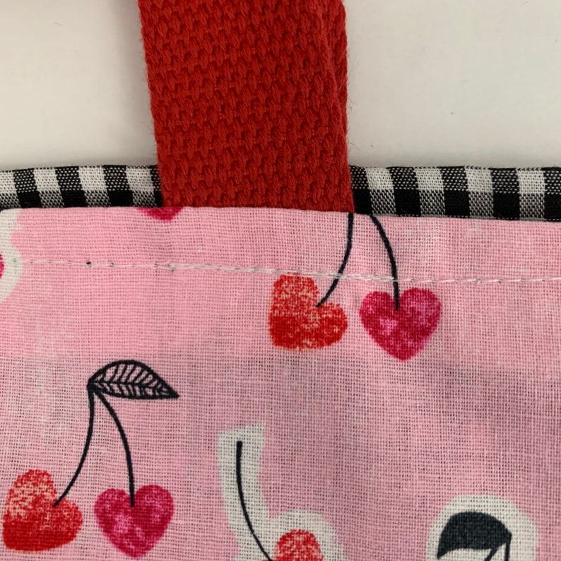 K&ouml;nnte beinhalten: Eine rosa Stofftasche mit einem roten Riemen und einem schwarz-wei&szlig; karierten Muster. Die Tasche hat ein Design aus roten Kirschen mit schwarzen Stielen und Bl&auml;ttern.