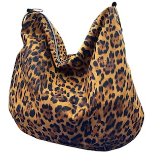 May include: A slouchy hobo bag featuring a bold leopard print pattern in shades of brown, tan, and black. The bag includes a top zipper closure and two circular metallic rings attached to the upper corners for the strap connection points.