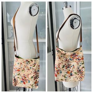Puede incluir: Un bolso bandolera beige con estampado floral y correa de cuero marr&oacute;n. El bolso tiene un forro verde.