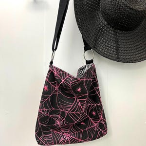 K&ouml;nnte beinhalten: Schwarze Tragetasche mit einem pinkfarbenen Spinnennetz- und Spinnenmuster. Die Tasche hat einen schwarzen Riemen und zwei silberne Ringe.