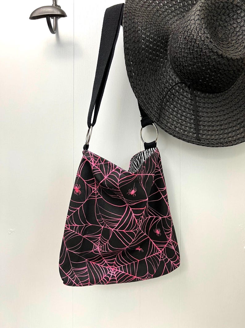 K&ouml;nnte beinhalten: Schwarze Tragetasche mit einem pinkfarbenen Spinnennetz- und Spinnenmuster. Die Tasche hat einen schwarzen Riemen und zwei silberne Ringe.