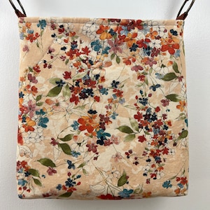Puede incluir: Una bolsa de tela beige con estampado floral con flores rojas, naranjas y azules. La bolsa tiene una correa de cuero marr&oacute;n y est&aacute; colgada de un gancho.