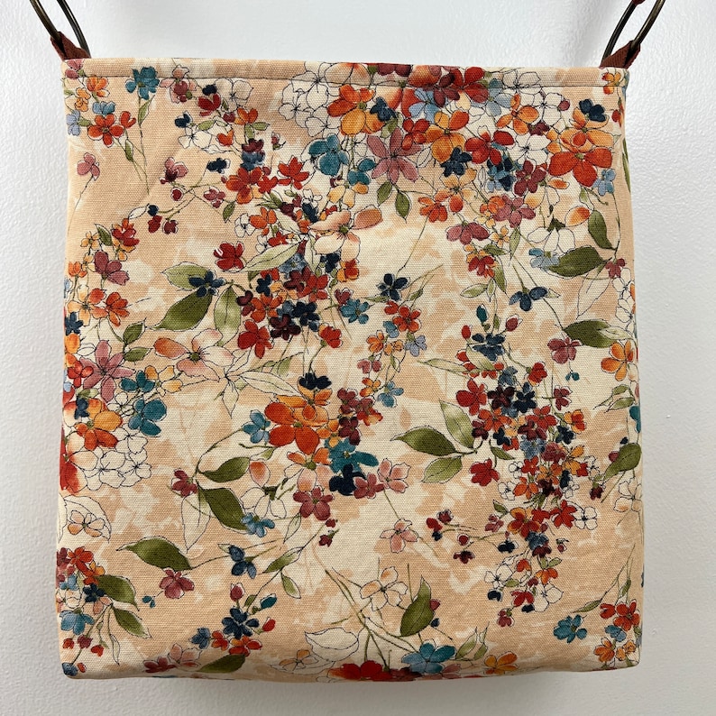 Puede incluir: Una bolsa de tela beige con estampado floral con flores rojas, naranjas y azules. La bolsa tiene una correa de cuero marr&oacute;n y est&aacute; colgada de un gancho.