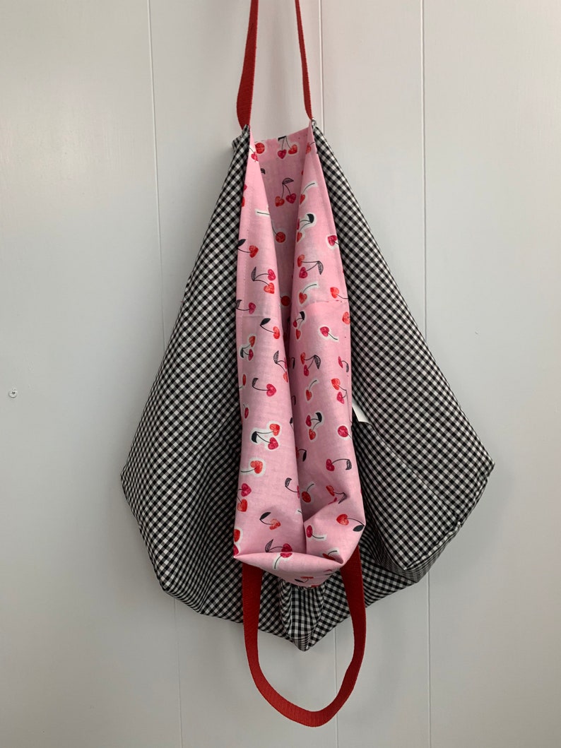 K&ouml;nnte beinhalten: Eine schwarze und wei&szlig;e Gingham-Tragetasche mit roten Griffen. Das Innere der Tasche ist mit einem rosa Stoff mit Kirschendruck ausgekleidet.