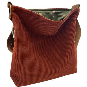 May include: A rust-colored corduroy shoulder bag with a brown strap and metal hardware. The interior lining is a dark green fabric with a whimsical mushroom pattern. The bag is open, revealing the patterned lining.