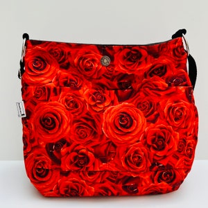 May include: A red floral print shoulder bag with a black strap and silver metal rings. The bag features a repeating pattern of red roses. It has two front pockets and a magnetic snap closure. A small tag is attached to the side.