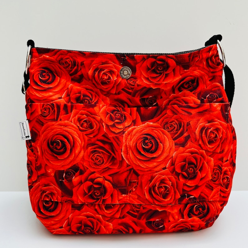 May include: A red floral print shoulder bag with a black strap and silver metal rings. The bag features a repeating pattern of red roses. It has two front pockets and a magnetic snap closure. A small tag is attached to the side.