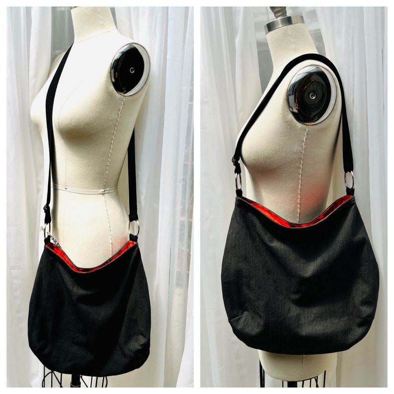 May include: Black corduroy shoulder bag with a red interior. The bag has a long black strap and silver-colored metal rings. The bag is displayed on a mannequin against a white curtain backdrop.
