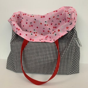 K&ouml;nnte beinhalten: Eine schwarz-wei&szlig;e Gingham-Tragetasche mit roten Griffen. Das Innere der Tasche ist mit pinkfarbenem Stoff mit einem Kirschmuster ausgekleidet. Die Tasche hat ein Etikett mit der Aufschrift "Handgefertigt".