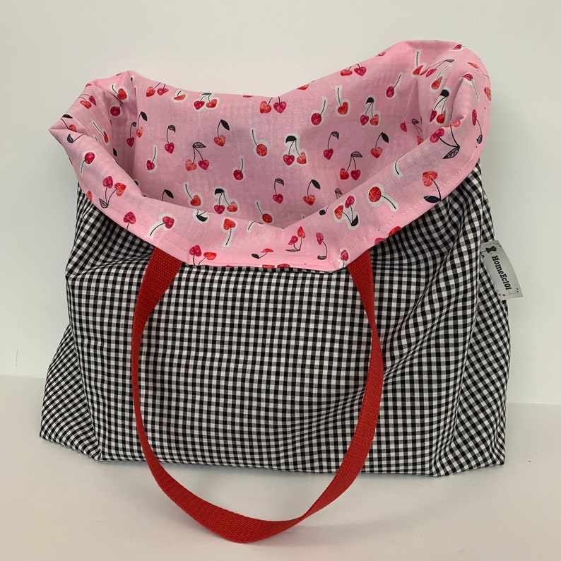 K&ouml;nnte beinhalten: Eine schwarz-wei&szlig;e Gingham-Tragetasche mit roten Griffen. Das Innere der Tasche ist mit pinkfarbenem Stoff mit einem Kirschmuster ausgekleidet. Die Tasche hat ein Etikett mit der Aufschrift "Handgefertigt".