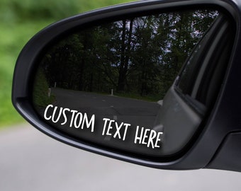 May include: A black car side mirror with a white vinyl sticker that says "CUSTOM TEXT HERE".