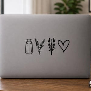 Op de afbeelding: Een matgrijze laptopdeksel met een zwarte vinylsticker. Het ontwerp bestaat uit vier afzonderlijke iconen die horizontaal zijn gerangschikt: een zoutvaatje, een takje rozemarijn, een lavendeltak en een handgetekende hartvorm.