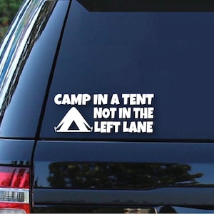 Pode incluir: Autocolante branco com o texto "CAMP IN A TENT NOT IN THE LEFT LANE" e um gráfico de tenda. O autocolante é aplicado numa janela escura de um veículo, sugerindo um tema de atividades ao ar livre ou campismo.