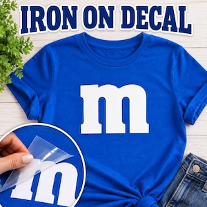 May include: A royal blue t-shirt featuring a large white lowercase letter m on the front. The text IRON ON DECAL appears at the top. A circular inset shows a hand peeling a clear plastic film away from a white letter m decal on blue fabric.