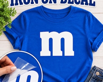 May include: A royal blue t-shirt featuring a large white lowercase letter m on the front. The text IRON ON DECAL appears at the top. A circular inset shows a hand peeling a clear plastic film away from a white letter m decal on blue fabric.