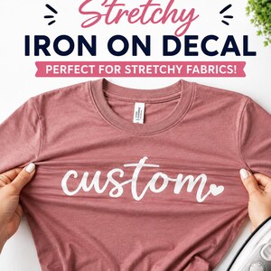 May include: A dusty rose-colored t-shirt featuring the white text "custom" in a cursive script with a small heart icon at the end. The shirt is being stretched horizontally to demonstrate the flexibility of the white iron-on decal material.