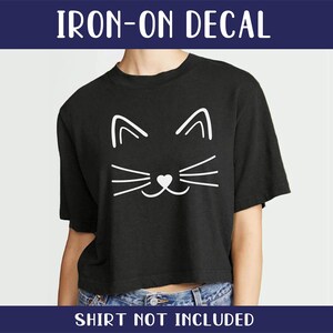 Op de afbeelding: Strijk-op sticker van een wit kattengezicht met een hartvormige neus op een zwart T-shirt. "IRON-ON DECAL" en "SHIRT NOT INCLUDED" staan in witte tekst op een blauwe achtergrond.