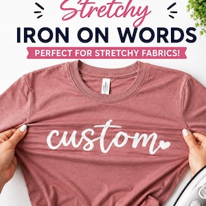 May include: A mauve crew neck t-shirt featuring the white script text custom with a small heart icon. The text Stretchy IRON ON WORDS PERFECT FOR STRETCHY FABRICS is displayed above the shirt. An iron is visible in the bottom right corner.