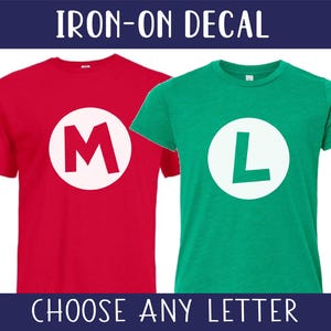 May include: Two short-sleeved t-shirts, one red and one green, each with a large white circle containing a letter. The red shirt has an "M", and the green shirt has an "L". The text "IRON-ON DECAL" and "CHOOSE ANY LETTER" are displayed.