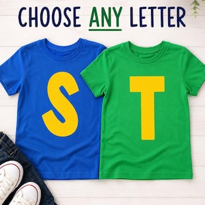 Pode incluir: Duas camisetas de manga curta, uma azul com uma letra S amarela grande e uma verde com uma letra T amarela grande. O texto CHOOSE ANY LETTER está impresso acima das camisetas. Calças jeans azul-escuras e tênis brancos são visíveis.