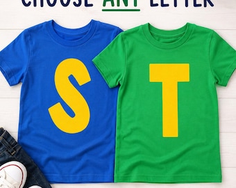May include: Two short-sleeve t-shirts, one blue with a large yellow letter S and one green with a large yellow letter T. The text CHOOSE ANY LETTER is printed above the shirts. Dark blue denim jeans and white canvas sneakers are visible below.