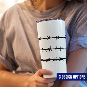 May include: A white tumbler with a black barbed wire design. The tumbler has a black lid and is held by a person wearing a gray t-shirt. The text "3 DESIGN OPTIONS" is in a blue rectangle at the bottom of the image.