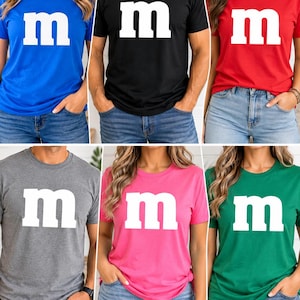 May include: A collection of six short-sleeved crew neck t-shirts in blue, black, red, gray, pink, and green. Each shirt features a large, white, lowercase letter m printed in the center of the chest, mimicking the iconic candy logo design.
